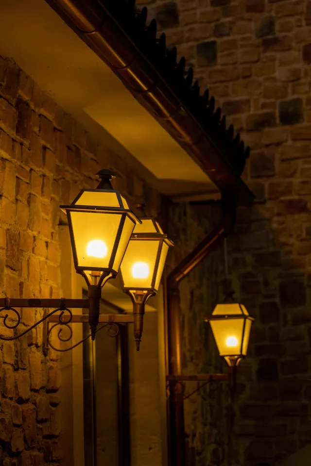 Three glowing, vintage-style lanterns mounted on a rustic brick wall under a dark wooden roof overhang at night.